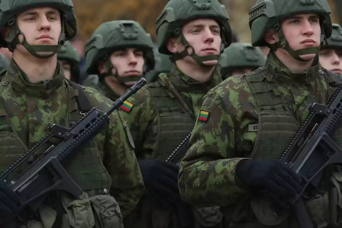 Soldados con uniforme de camuflaje y casco verde marchando en formación, portando rifles y con la bandera de Lituania en el brazo