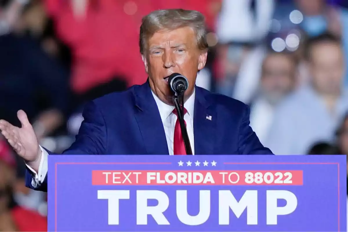 Hombre de cabello rubio con traje azul y corbata roja hablando en un mitin político detrás de un podio que tiene un cartel con el texto Trump y un mensaje para enviar un mensaje de texto a un número específico