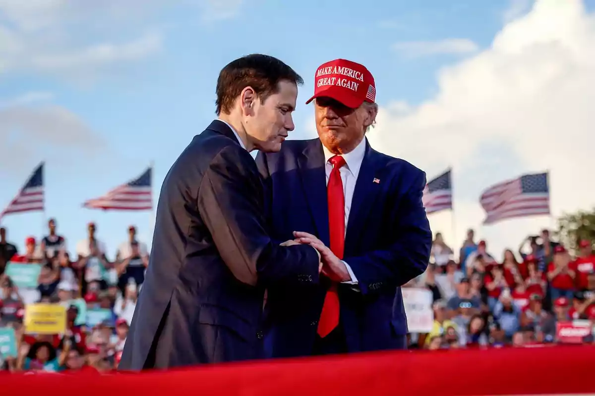Dos hombres con traje se dan la mano en un evento al aire libre con banderas de Estados Unidos y una multitud al fondo uno de ellos lleva una gorra roja Dos hombres con traje se dan la mano en un evento al aire libre con banderas de Estados Unidos y una multitud al fondo uno de ellos lleva una gorra roja