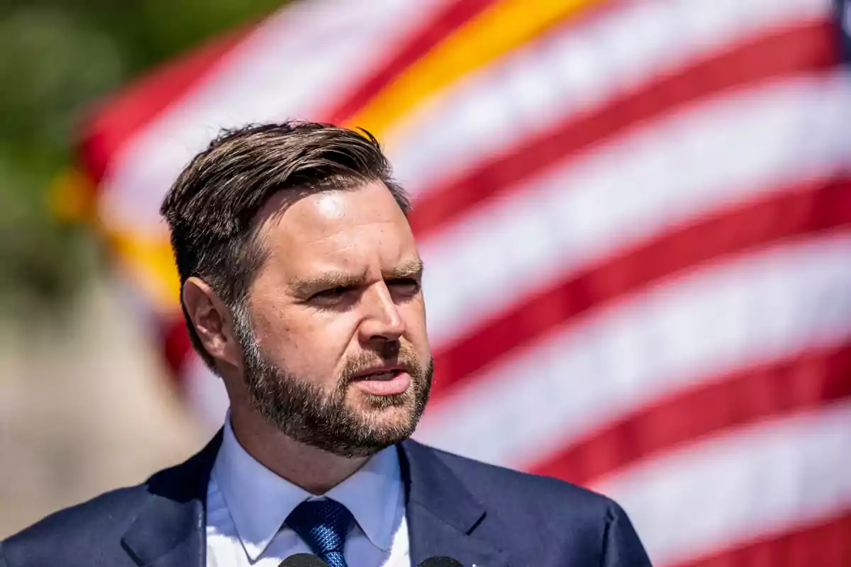 Man in a suit speaking in front of a blurred United States flag Man in a suit speaking in front of a blurred United States flag