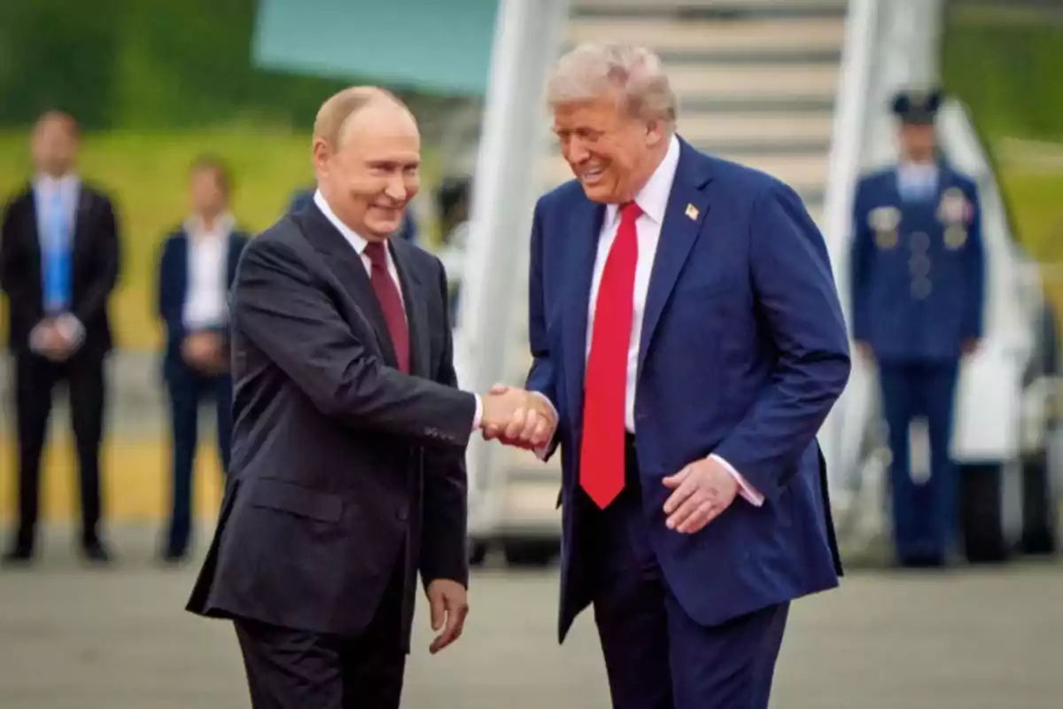 Two men in suits shake hands and smile while other people watch in the background in an outdoor setting. Two men in suits shake hands and smile while other people watch in the background in an outdoor setting.