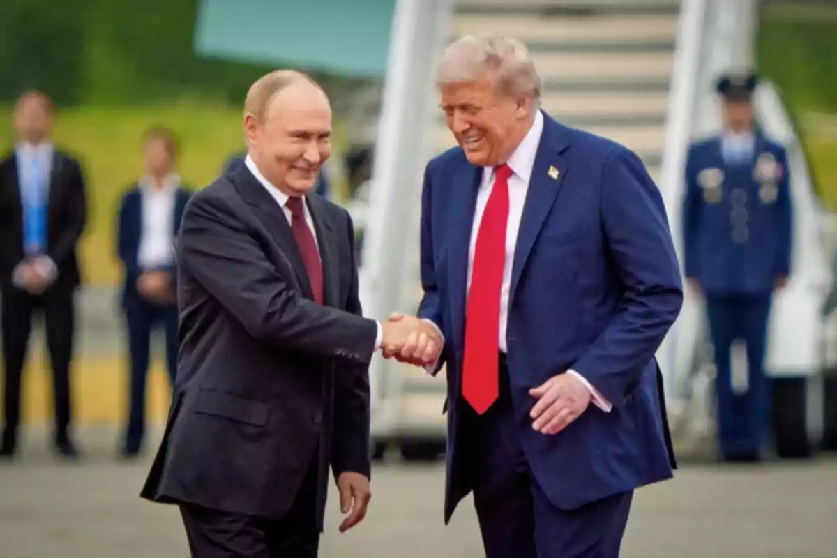 Two men in suits shake hands and smile in an outdoor setting with uniformed people in the background. Two men in suits shake hands and smile in an outdoor setting with uniformed people in the background.