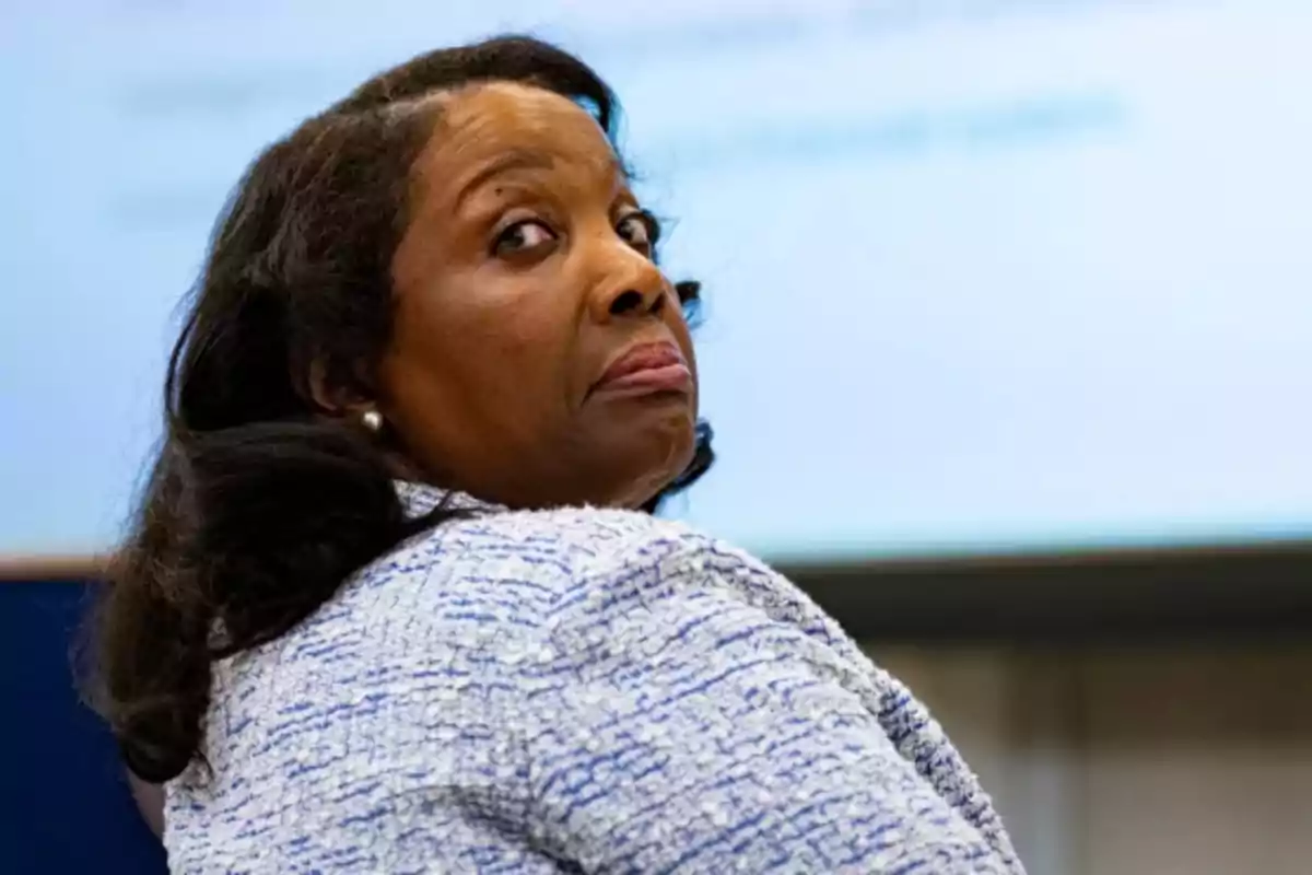 Dark-haired woman with loose hair wearing a light jacket looking back with a serious expression