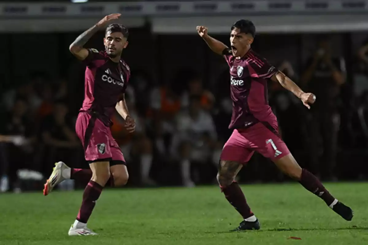 Dos futbolistas con uniforme color vino celebran un gol en la cancha durante un partido nocturno