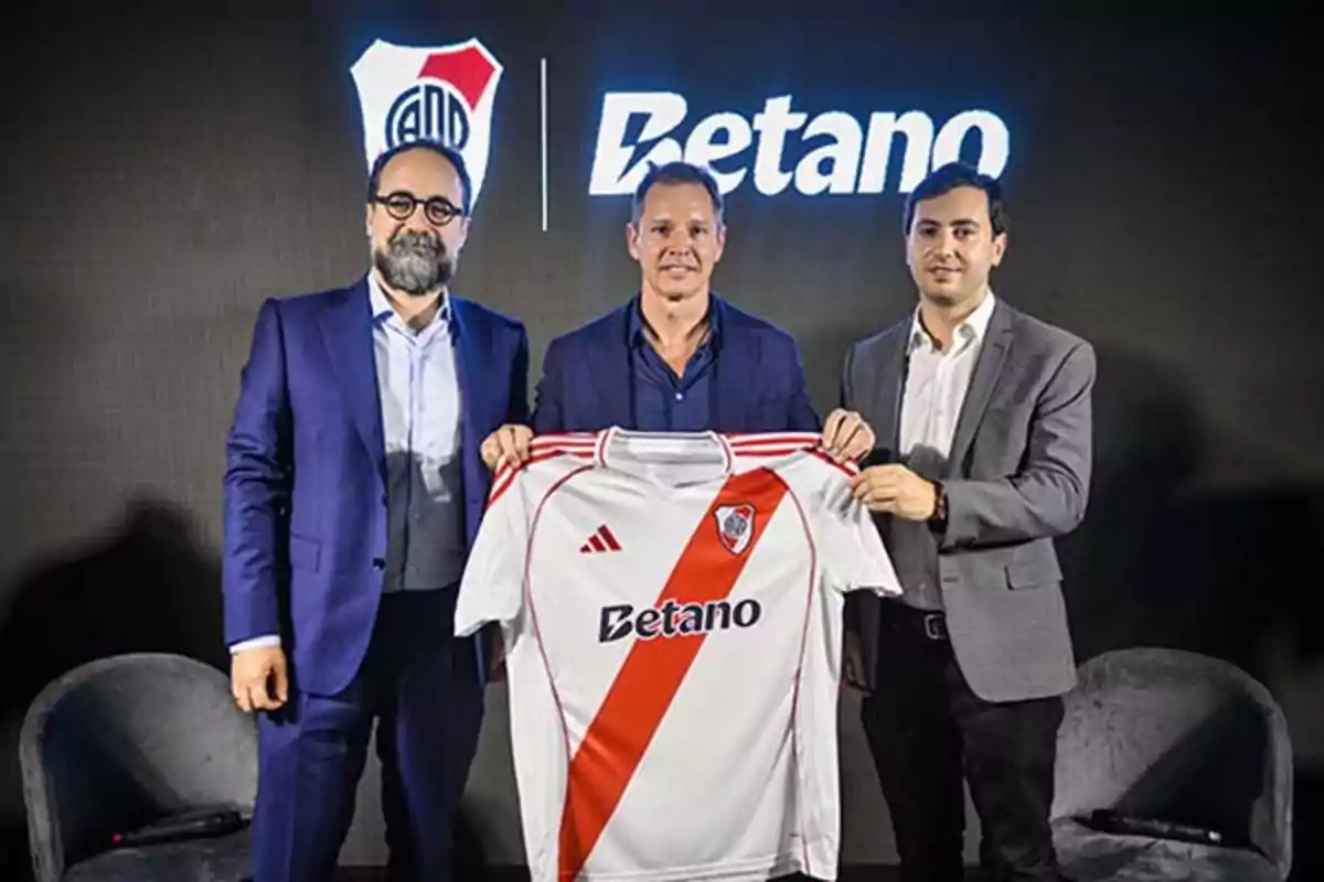 Three men in suits pose holding a River Plate jersey with the Betano logo at a launch event. Three men in suits pose holding a River Plate jersey with the Betano logo at a launch event.