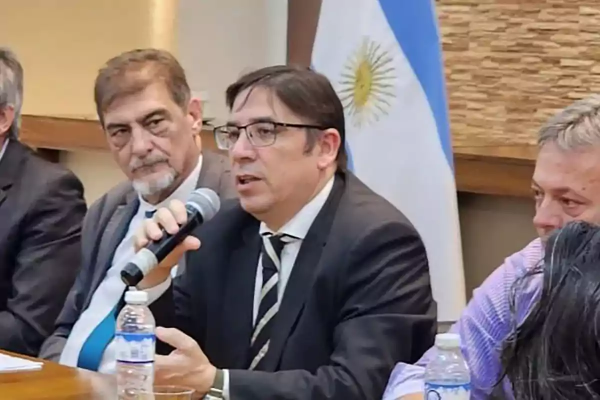 A man wearing glasses and a suit speaks into a microphone at a meeting table with other people, with an Argentine flag visible in the background. A man wearing glasses and a suit speaks into a microphone at a meeting table with other people, with an Argentine flag visible in the background.