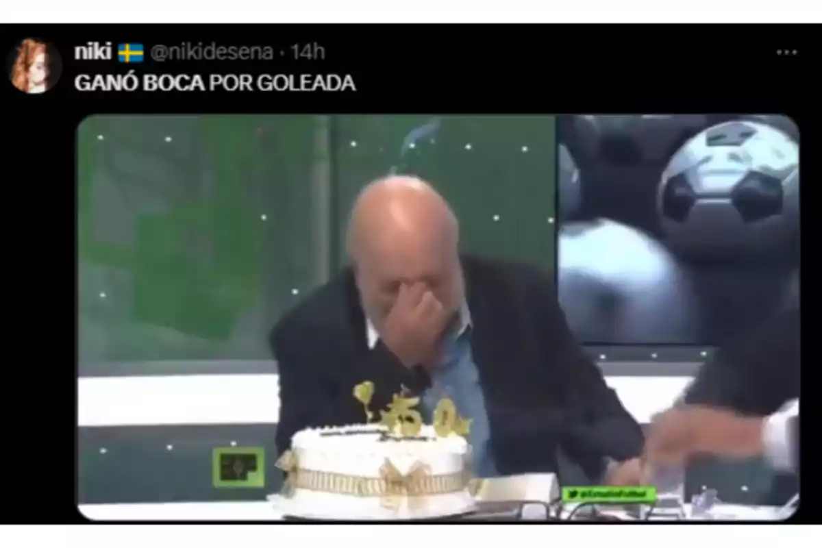 Bald man with a beard covering his face with his hand while sitting in front of a white cake decorated with the number 50 on a soccer-themed television set Bald man with a beard covering his face with his hand while sitting in front of a white cake decorated with the number 50 on a soccer-themed television set