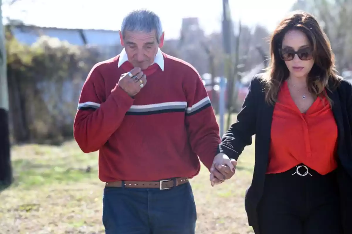 Un hombre mayor con suéter rojo y una mujer con blusa roja y gafas de sol caminan tomados de la mano en un entorno al aire libre Un hombre mayor con suéter rojo y una mujer con blusa roja y gafas de sol caminan tomados de la mano en un entorno al aire libre