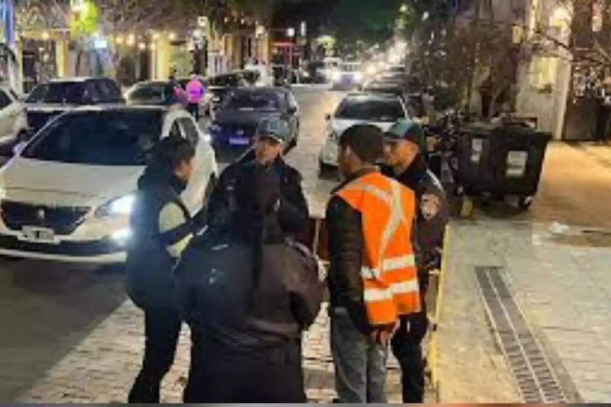 Several people, including police officers and a worker wearing a reflective vest, are talking on a street lit up at night with parked cars and storefront lights in the background. Several people, including police officers and a worker wearing a reflective vest, are talking on a street lit up at night with parked cars and storefront lights in the background.
