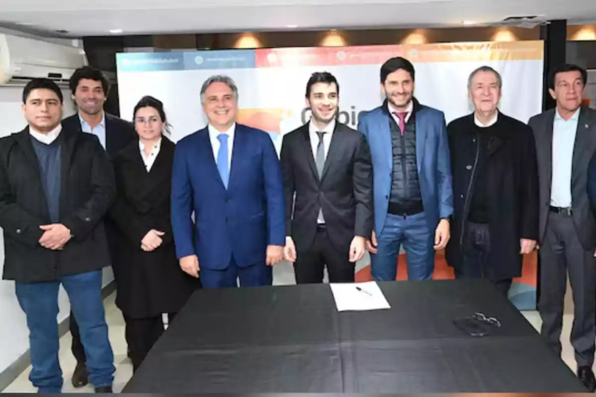 Ocho personas posan de pie y sonríen frente a una mesa cubierta con un mantel negro en un ambiente de oficina Ocho personas posan de pie y sonríen frente a una mesa cubierta con un mantel negro en un ambiente de oficina