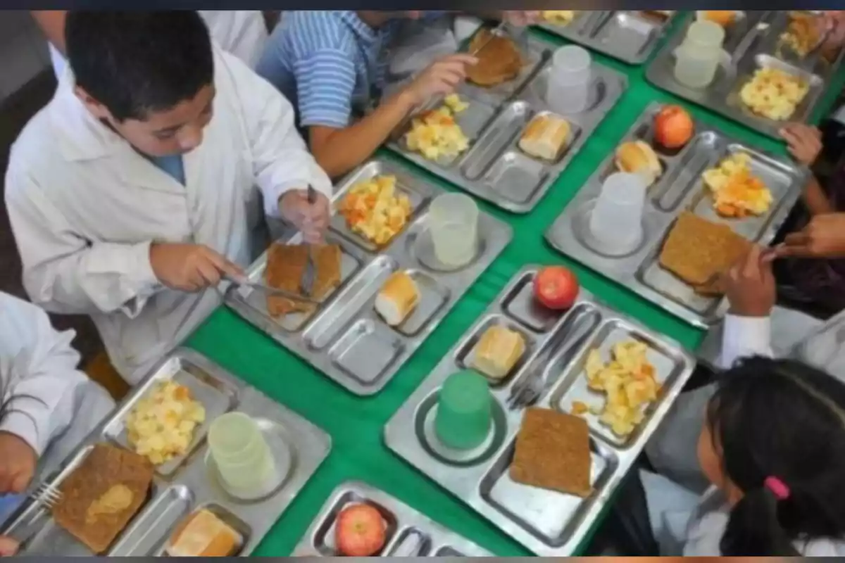 Niños sentados en una mesa comiendo en una cafetería escolar con bandejas de comida que incluyen pan, una porción de guiso, una manzana y un vaso de plástico Niños sentados en una mesa comiendo en una cafetería escolar con bandejas de comida que incluyen pan, una porción de guiso, una manzana y un vaso de plástico