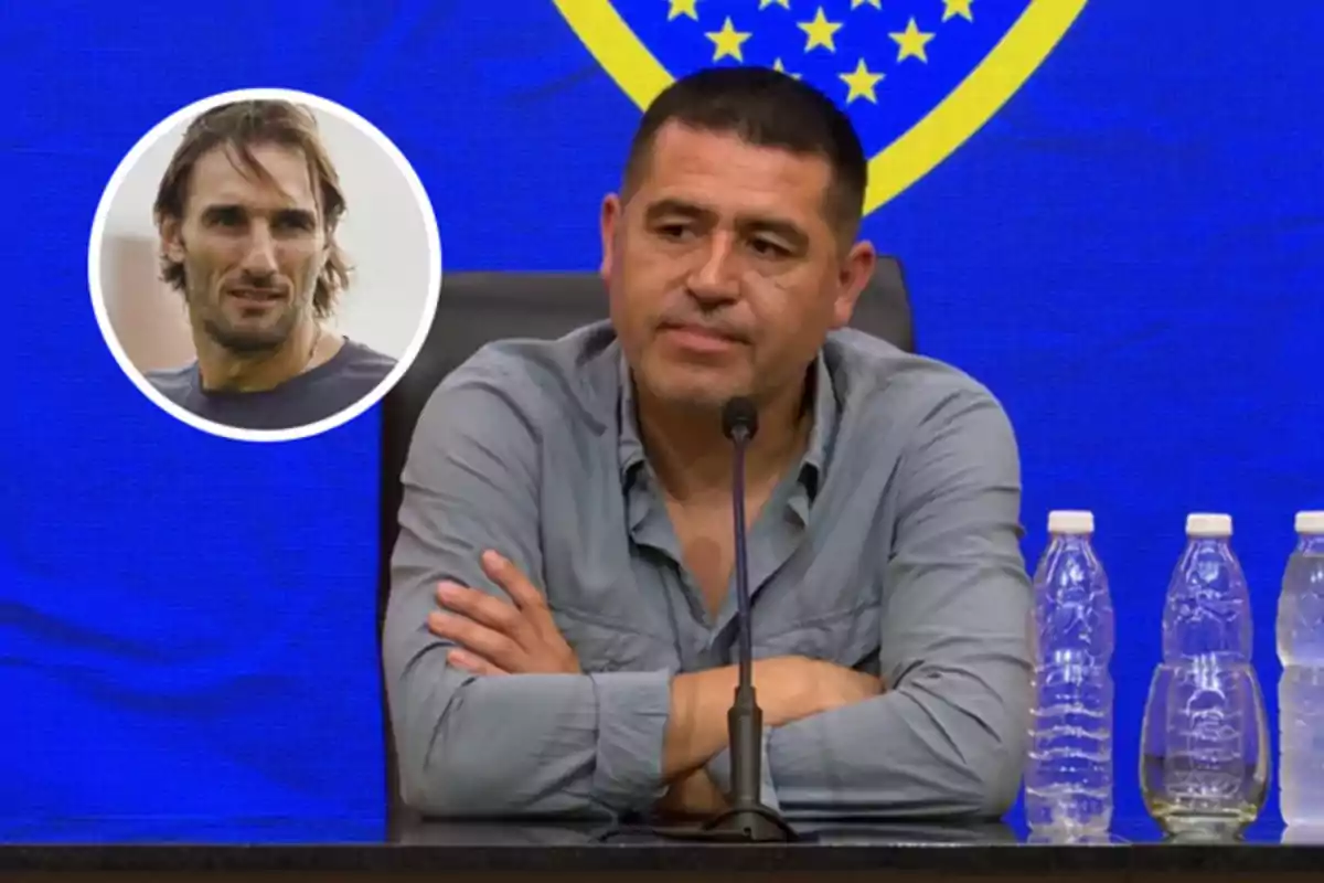 Hombre de cabello corto y camisa gris sentado en una conferencia de prensa con fondo azul y amarillo, varias botellas de agua sobre la mesa y un recuadro con el rostro de otro hombre en la esquina izquierda