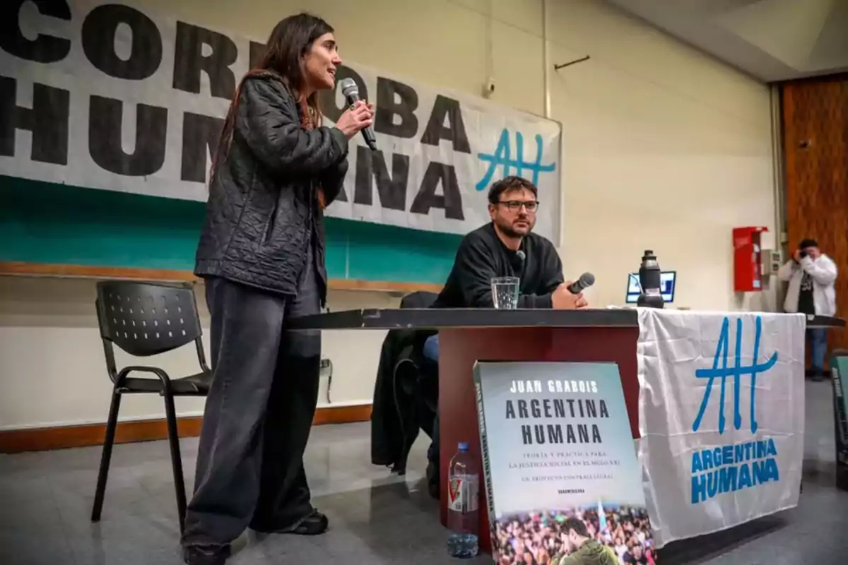 Una mujer de pie habla por micrófono junto a un hombre sentado en una mesa durante una presentación de un libro titulado Argentina Humana en un auditorio decorado con carteles y una bandera del mismo nombre Una mujer de pie habla por micrófono junto a un hombre sentado en una mesa durante una presentación de un libro titulado Argentina Humana en un auditorio decorado con carteles y una bandera del mismo nombre