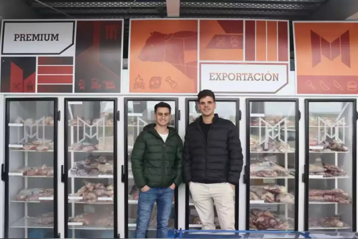 Dos hombres jóvenes posan frente a vitrinas de carne en una tienda con letreros que dicen premium y exportación Dos hombres jóvenes posan frente a vitrinas de carne en una tienda con letreros que dicen premium y exportación