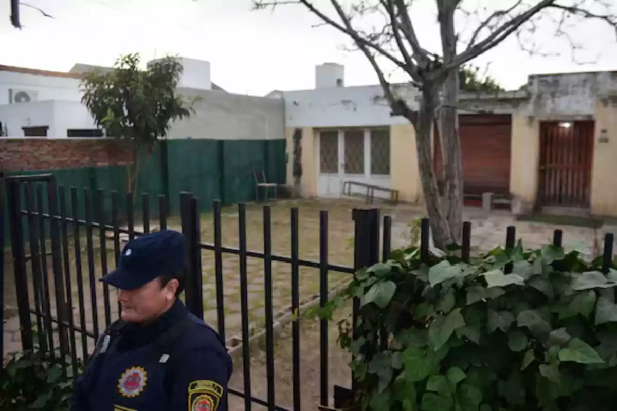 Una mujer policía custodia la entrada de una casa con jardín y rejas negras en un barrio residencial Una mujer policía custodia la entrada de una casa con jardín y rejas negras en un barrio residencial