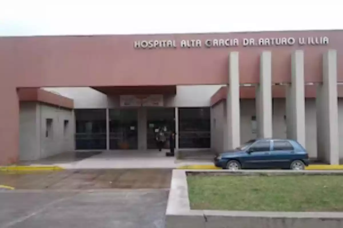 Fachada de un hospital con un auto azul estacionado al frente y una persona entrando al edificio