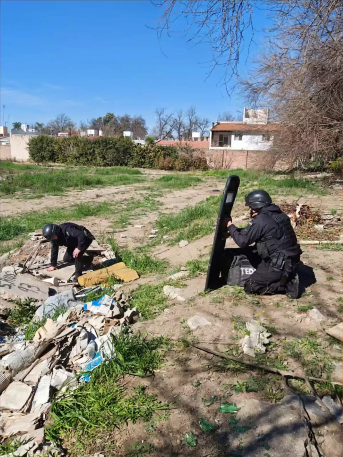 Dos personas con uniforme y casco de protección trabajan en un terreno baldío con escombros y pasto, una de ellas sostiene un escudo y la otra revisa el suelo
