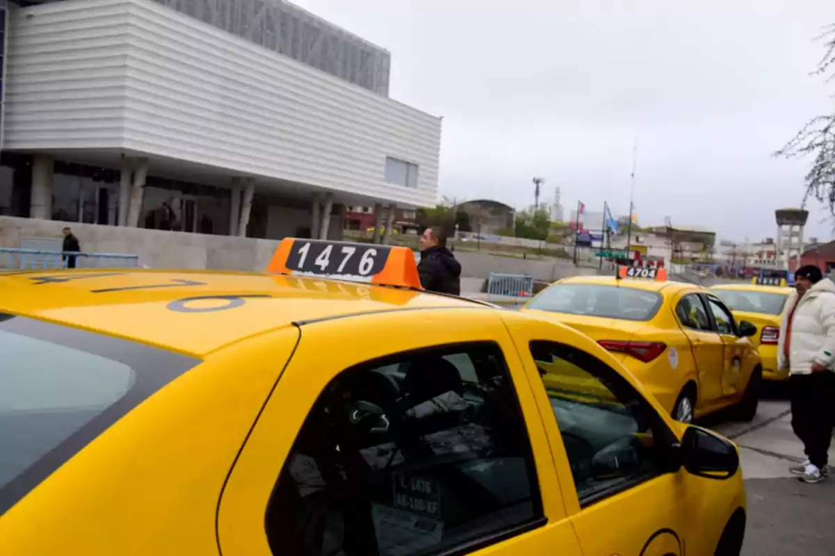 Varios taxis amarillos estacionados en fila frente a un edificio moderno mientras algunas personas caminan cerca