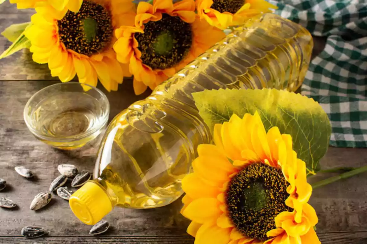 Botella de aceite de girasol junto a flores de girasol, semillas y un recipiente pequeño sobre una mesa de madera Botella de aceite de girasol junto a flores de girasol, semillas y un recipiente pequeño sobre una mesa de madera
