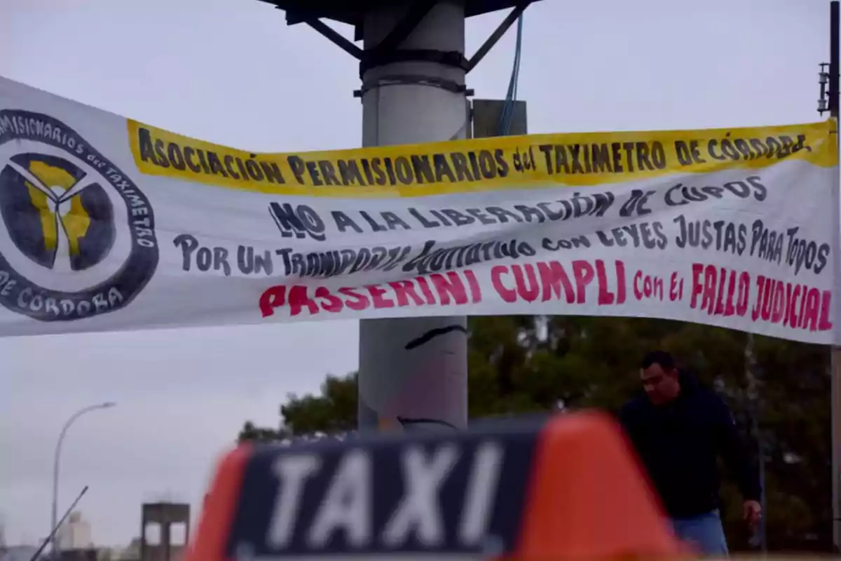 Pancarta de la Asociación Permisionarios del Taxímetro de Córdoba colgada en la vía pública con mensajes en contra de la liberación de cupos y exigiendo el cumplimiento de un fallo judicial, con un taxi y una persona en el fondo. Pancarta de la Asociación Permisionarios del Taxímetro de Córdoba colgada en la vía pública con mensajes en contra de la liberación de cupos y exigiendo el cumplimiento de un fallo judicial, con un taxi y una persona en el fondo.