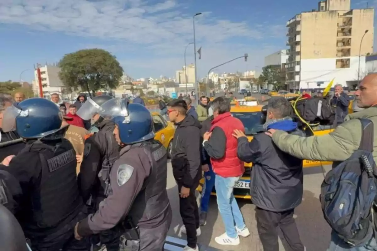 A group of police officers wearing helmets and carrying shields confronts several people in front of yellow taxis on a city street during a protest. A group of police officers wearing helmets and carrying shields confronts several people in front of yellow taxis on a city street during a protest.
