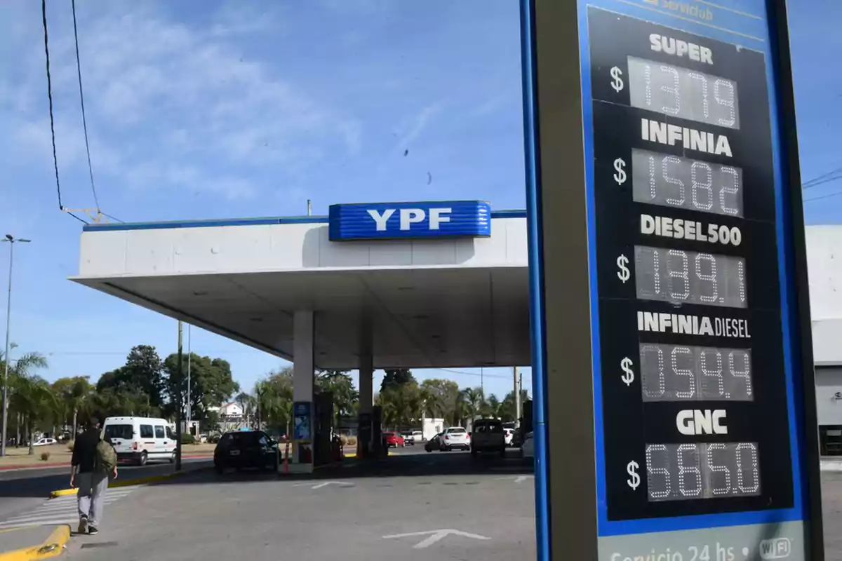 Estación de servicio YPF con cartel de precios de combustibles y autos cargando nafta en un día soleado.