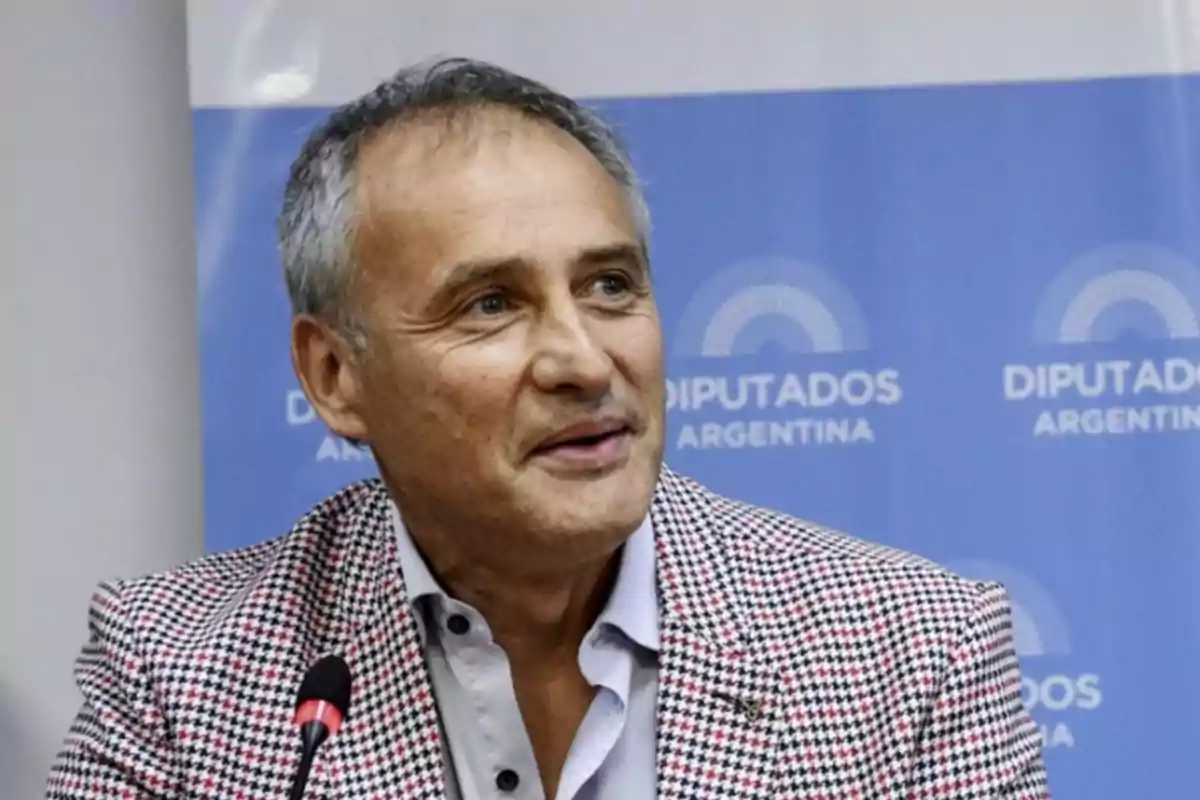 Gray-haired man in a plaid jacket speaking into a microphone with a blue background that says Deputies Argentina Gray-haired man in a plaid jacket speaking into a microphone with a blue background that says Deputies Argentina