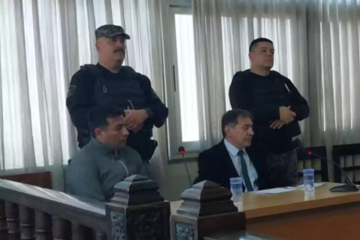 Two men sitting in a courtroom with two security guards standing behind them Two men sitting in a courtroom with two security guards standing behind them