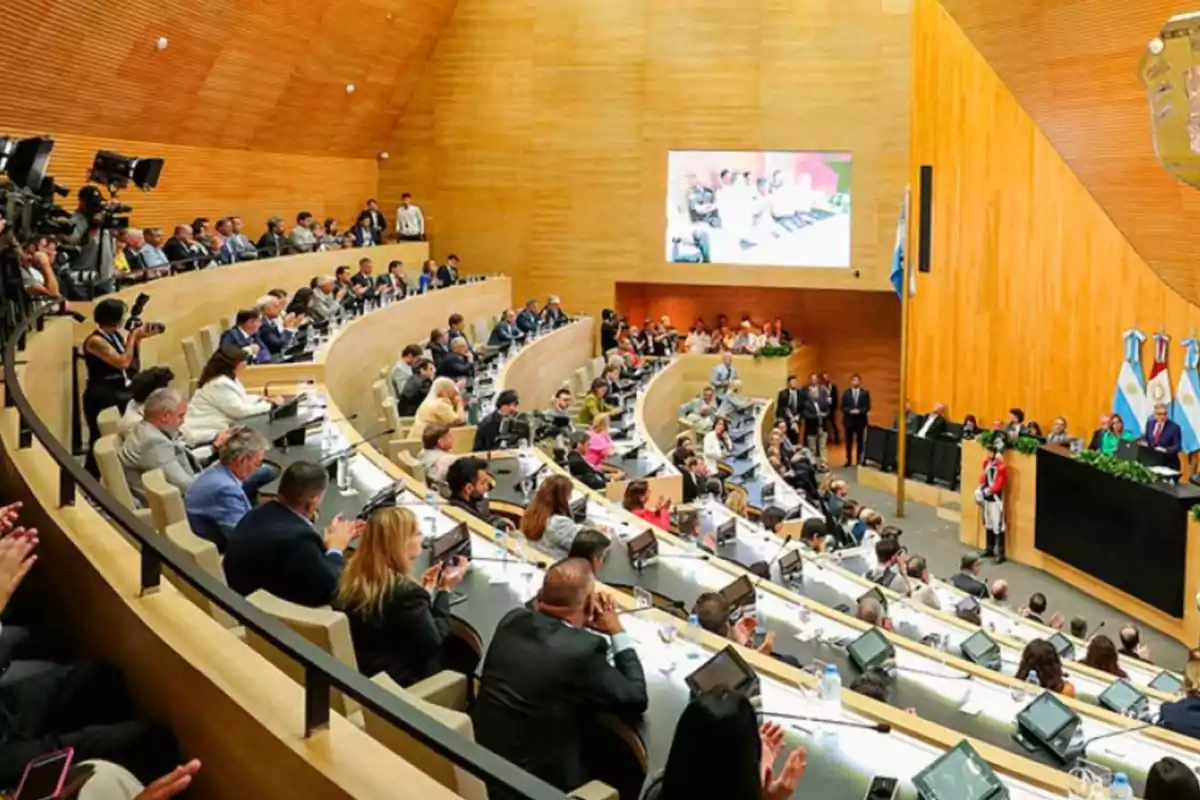 People gathered in a modern legislative chamber with wooden walls, several rows of seats, and screens, while some people applaud and others watch a presentation on the main stage. People gathered in a modern legislative chamber with wooden walls, several rows of seats, and screens, while some people applaud and others watch a presentation on the main stage.