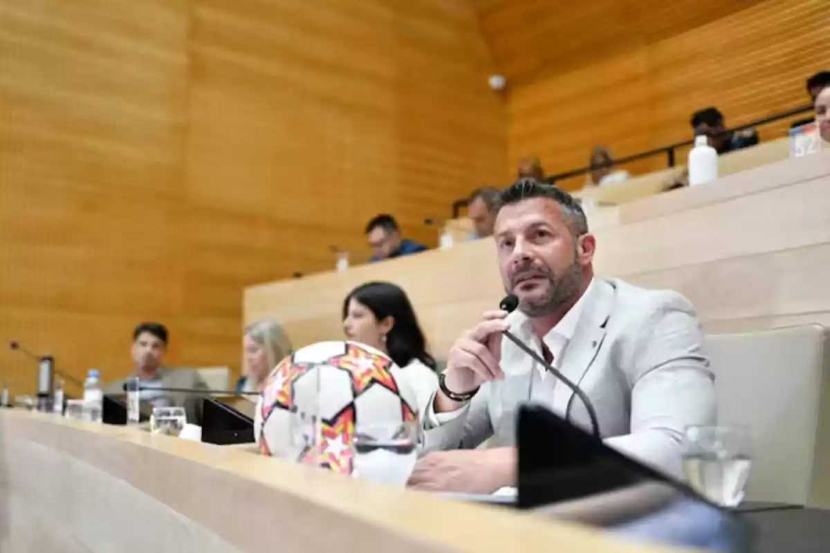 Man in a light-colored jacket speaking into a microphone in a conference room with a soccer ball on the table and several people in the background