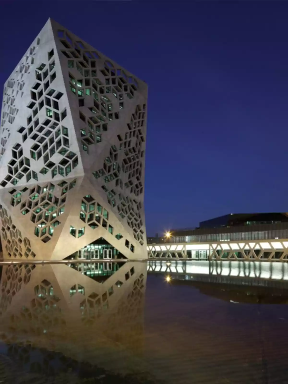 Edificio moderno con fachada geométrica iluminado por la noche y reflejado en un estanque de agua Edificio moderno con fachada geométrica iluminado por la noche y reflejado en un estanque de agua