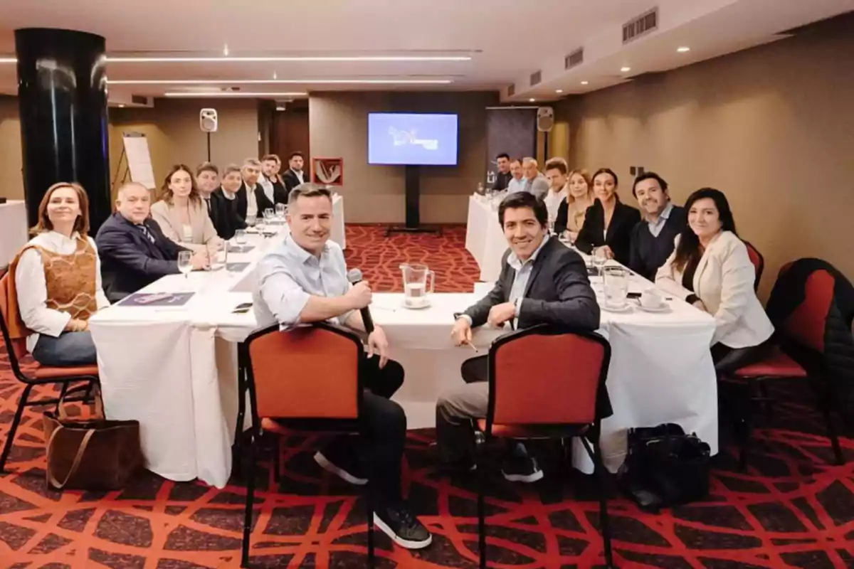 A group of people sitting around a table in a meeting room, with a screen in the background displaying a presentation.
