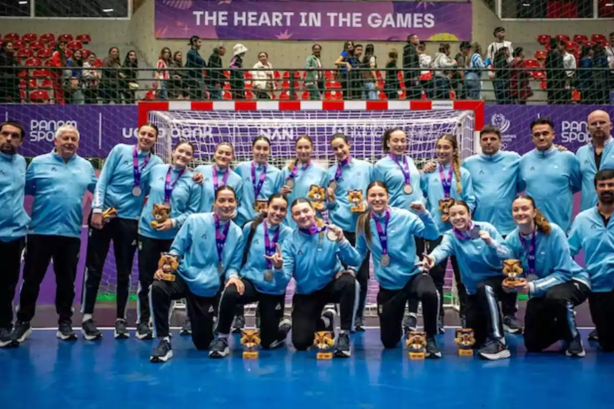 Equipo femenino de handball posando sonriente con medallas y trofeos frente a una portería en un estadio con público al fondo Equipo femenino de handball posando sonriente con medallas y trofeos frente a una portería en un estadio con público al fondo