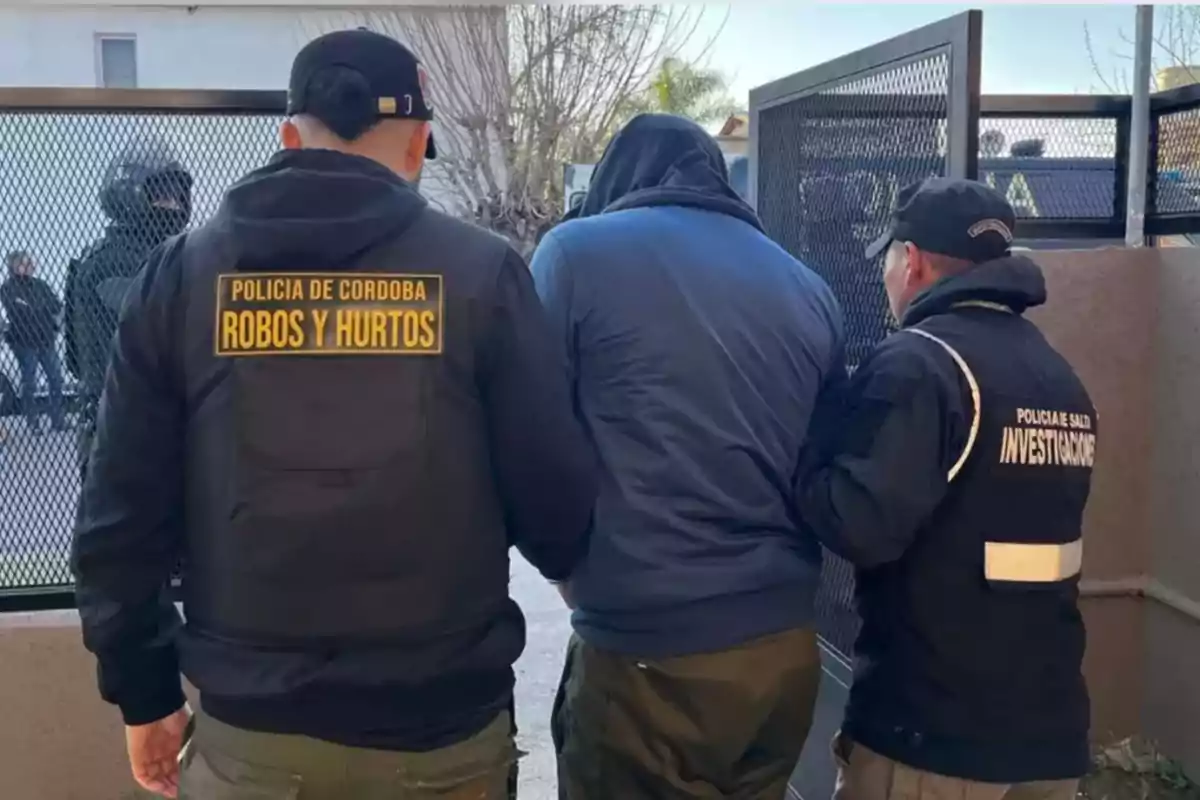 Tres agentes de policía escoltan a una persona con capucha hacia una puerta metálica Tres agentes de policía escoltan a una persona con capucha hacia una puerta metálica
