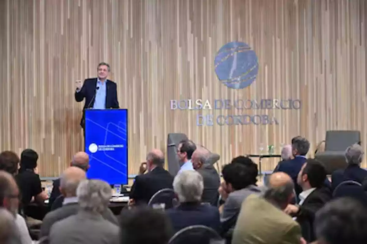 A man in a suit speaks at a blue podium in front of an audience in an auditorium with the Córdoba Stock Exchange logo on the back wall. A man in a suit speaks at a blue podium in front of an audience in an auditorium with the Córdoba Stock Exchange logo on the back wall.