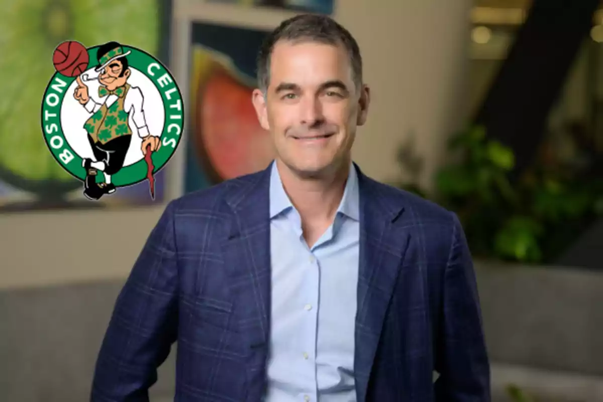 Hombre de cabello corto y canoso con saco azul posando sonriente frente al logo de los Boston Celtics