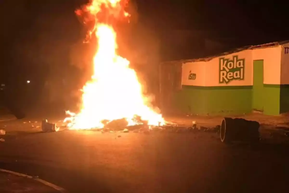 Fuego intenso en medio de una calle durante la noche con un edificio de Kola Real al fondo Fuego intenso en medio de una calle durante la noche con un edificio de Kola Real al fondo