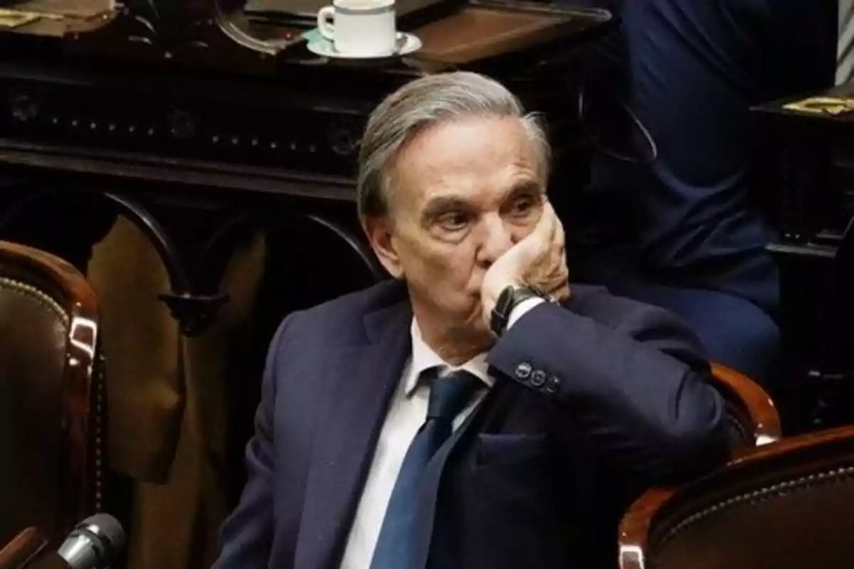 Older man in a blue suit sitting in a room, resting his head on his hand with a thoughtful expression