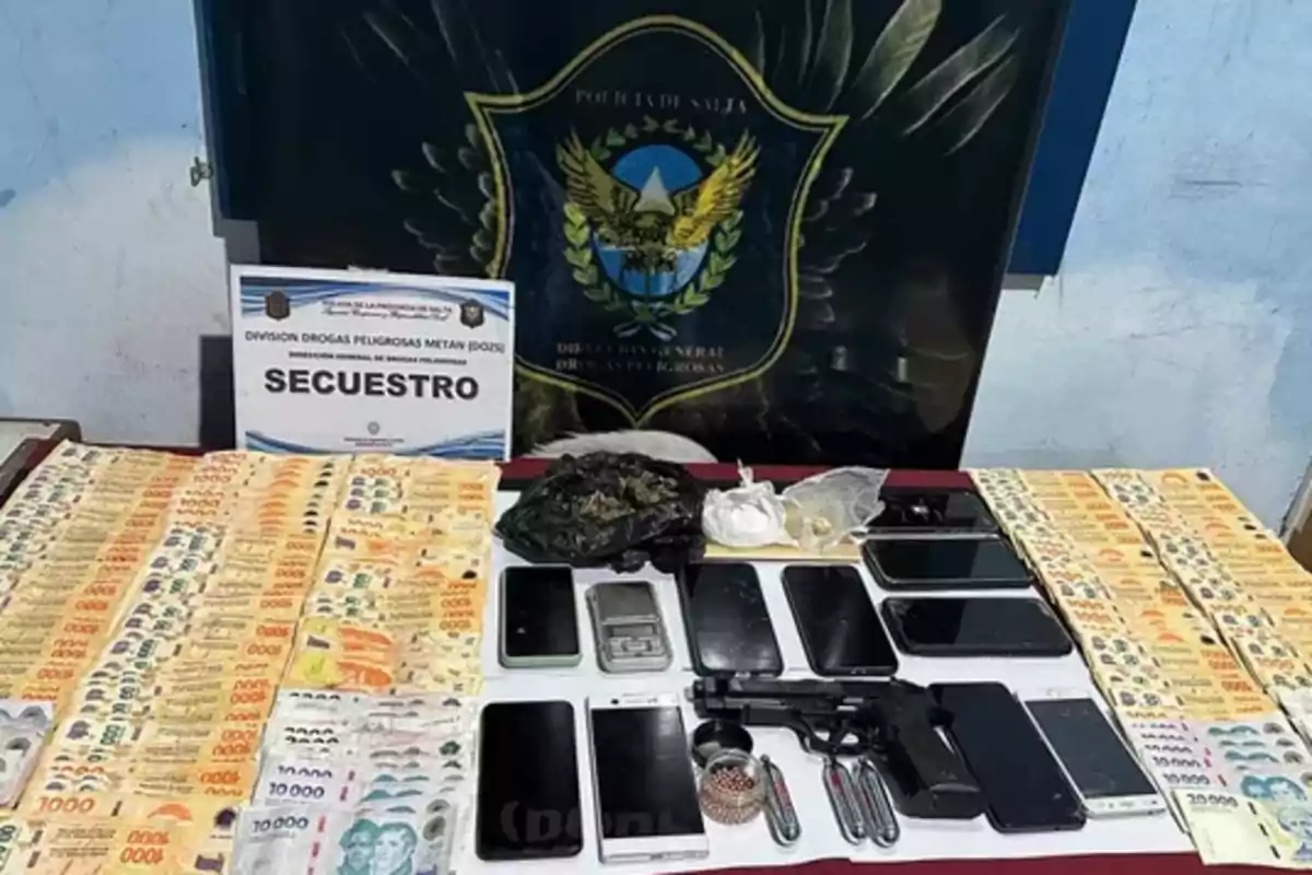 Billetes de diferentes denominaciones, varios teléfonos móviles, una balanza, una pistola, bolsas con sustancias y objetos metálicos exhibidos sobre una mesa frente a un cartel de la policía y un escudo oficial