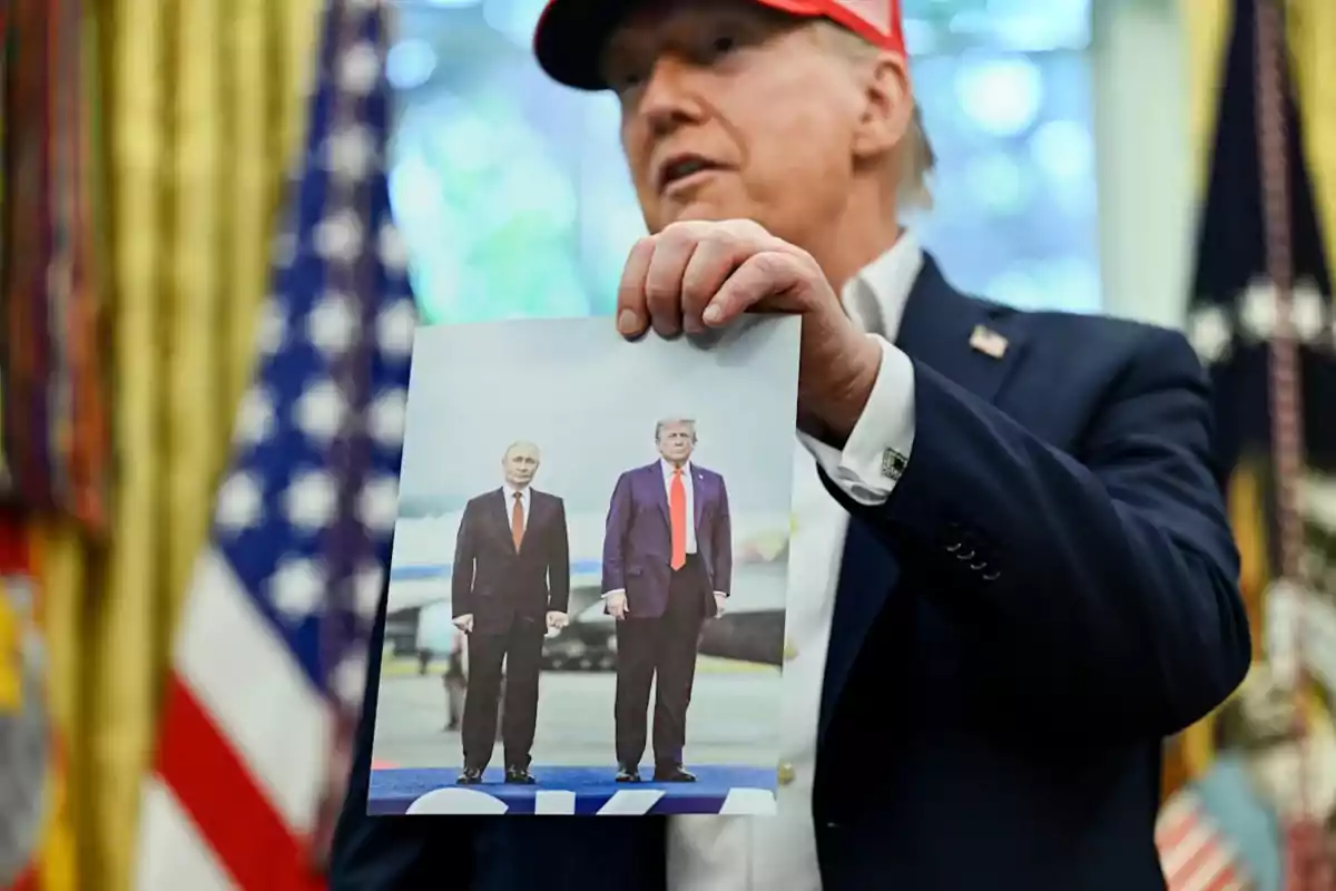 A man wearing a red cap is holding a photograph showing two people standing in formal suits and red ties in front of an outdoor background.
