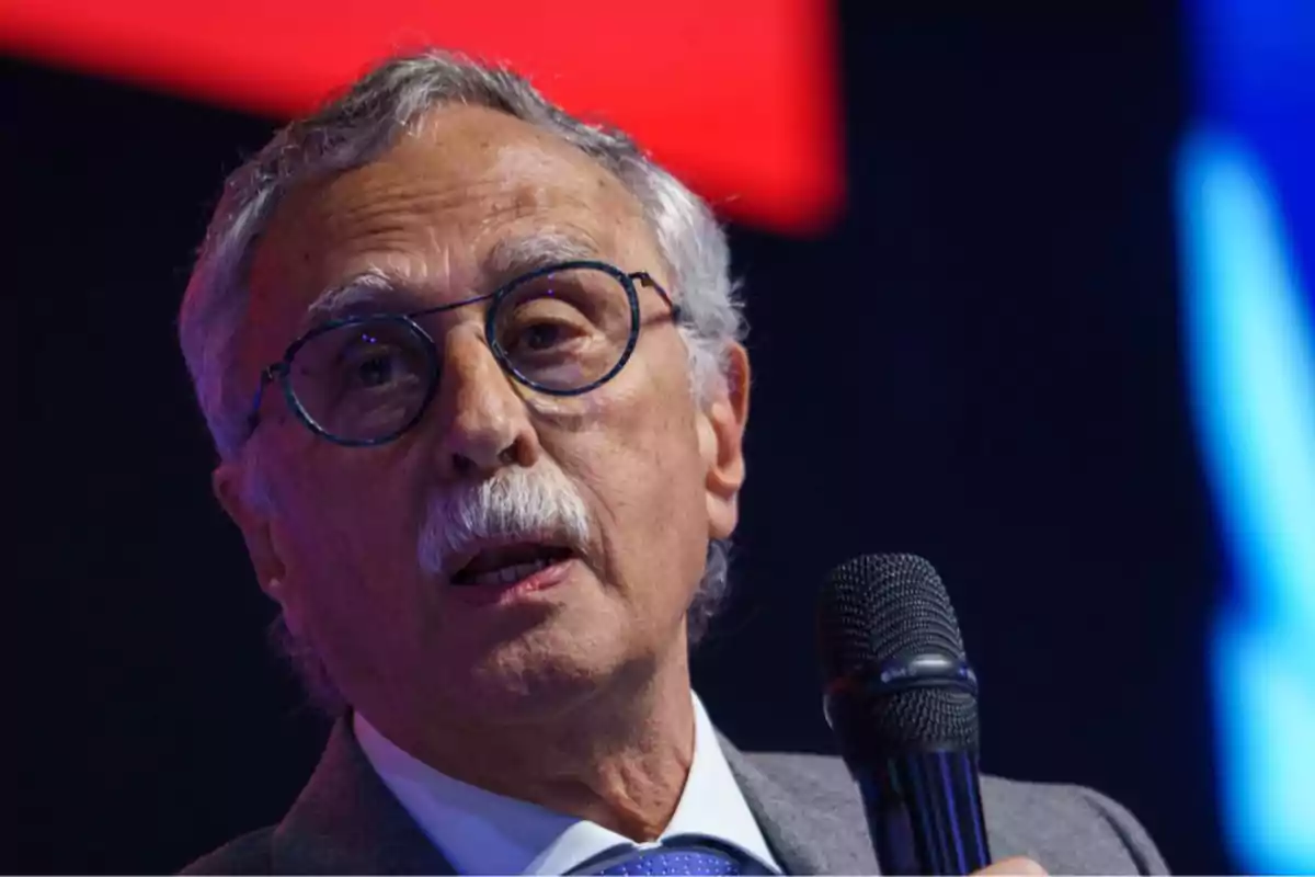 Older man with glasses and a mustache holding a microphone while speaking at an event