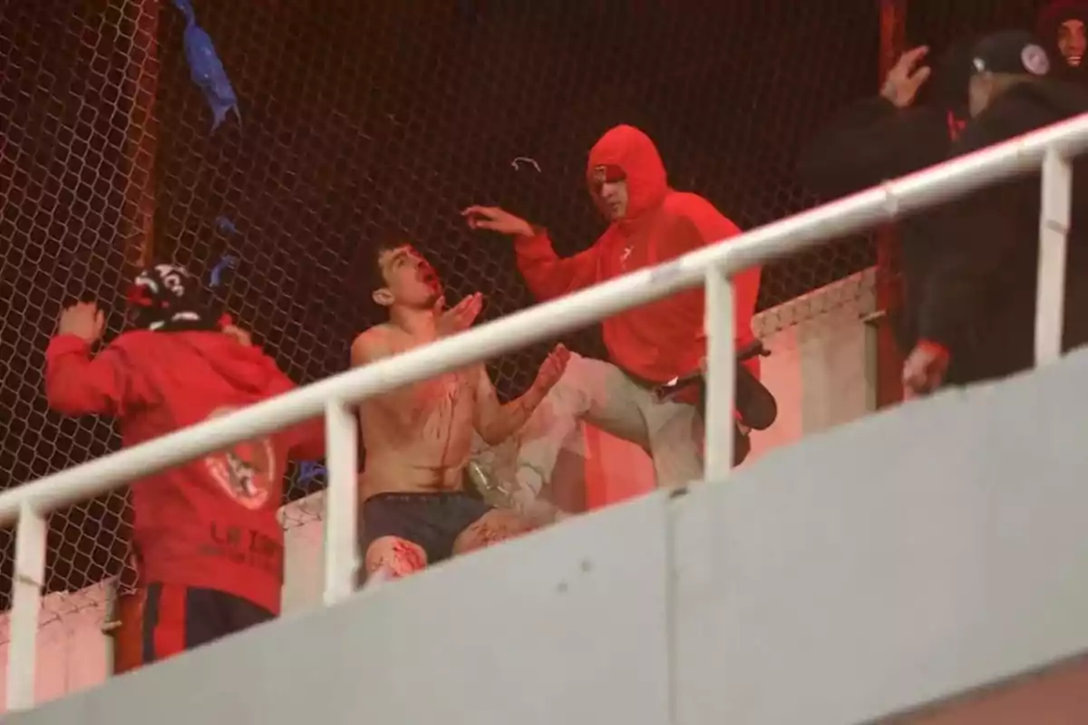 Un grupo de personas con ropa deportiva y capuchas rodea a un hombre sin camiseta y con sangre en el cuerpo junto a una reja en un estadio Un grupo de personas con ropa deportiva y capuchas rodea a un hombre sin camiseta y con sangre en el cuerpo junto a una reja en un estadio