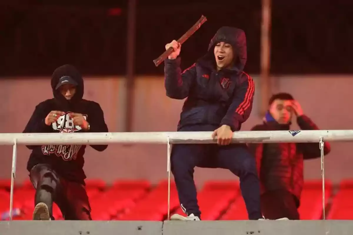 Tres jóvenes con ropa deportiva y capuchas están en una grada de estadio, uno de ellos sostiene un objeto de madera y parece estar gritando, mientras los otros dos observan o se preparan Tres jóvenes con ropa deportiva y capuchas están en una grada de estadio, uno de ellos sostiene un objeto de madera y parece estar gritando, mientras los otros dos observan o se preparan