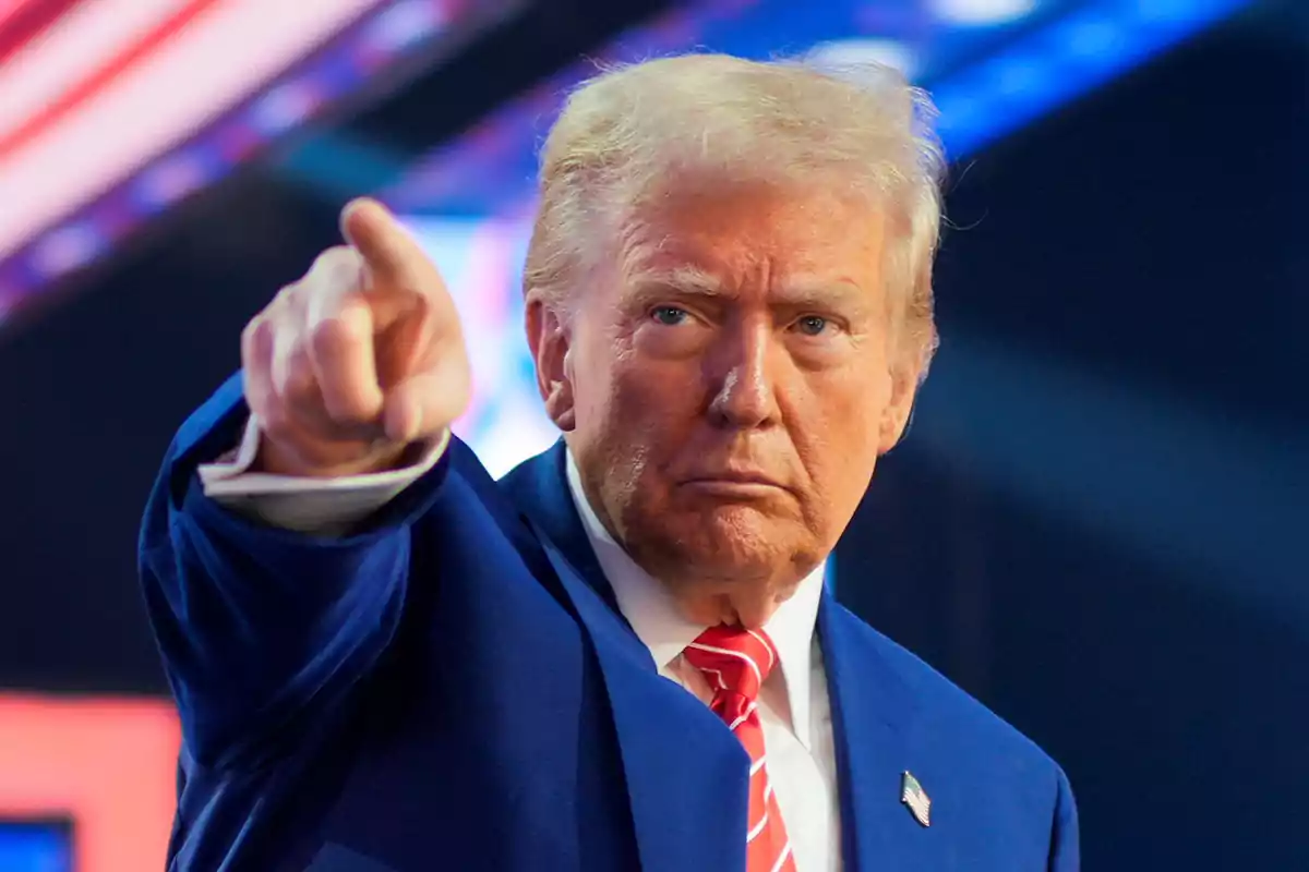 Hombre de cabello rubio con traje azul y corbata roja señalando hacia el frente con expresión seria