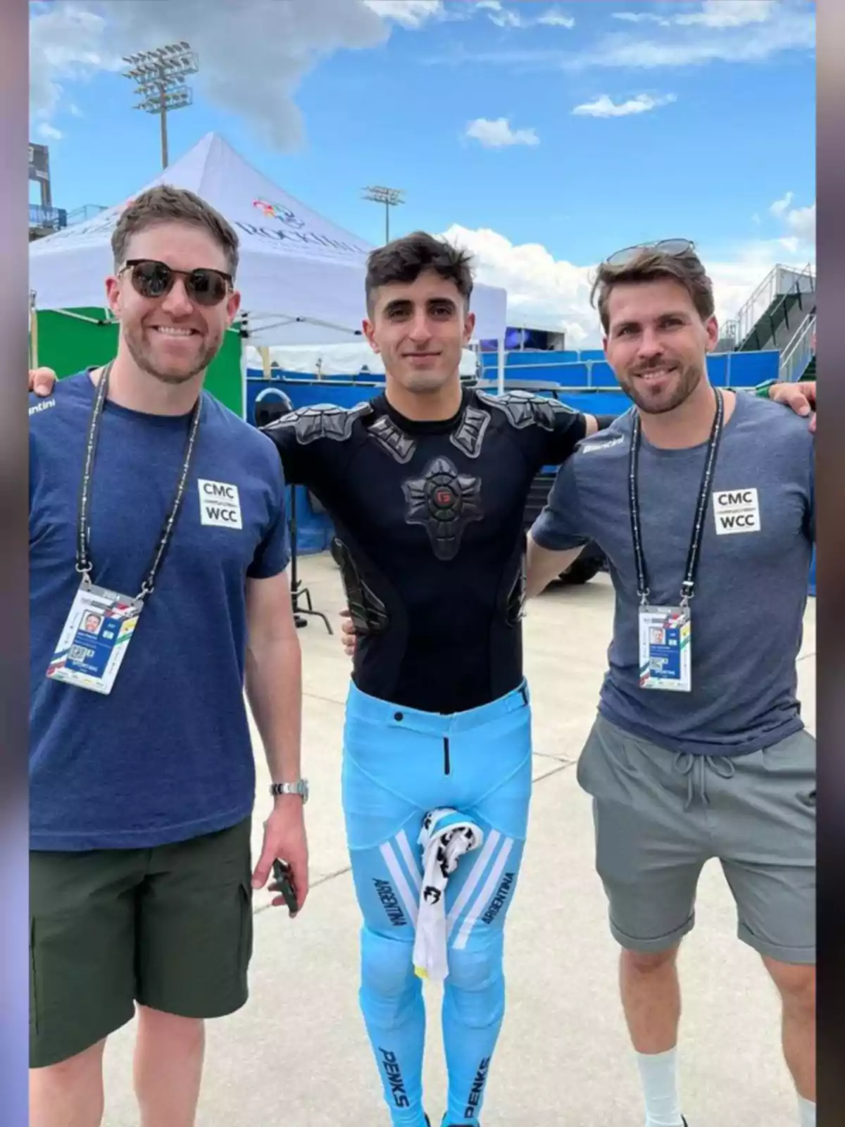 Three men pose smiling outdoors, one of them wearing a blue sports uniform with the word Argentina and protective gear, while the other two are dressed in casual clothes and badges, with a white tent and blue sky in the background. Three men pose smiling outdoors, one of them wearing a blue sports uniform with the word Argentina and protective gear, while the other two are dressed in casual clothes and badges, with a white tent and blue sky in the background.