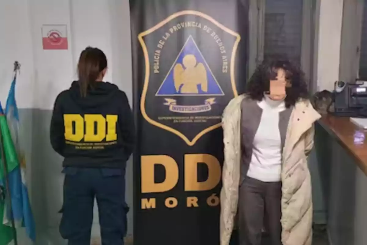 Una agente de policía de espaldas y una mujer esposada de frente están junto a un cartel de la DDI Morón en una comisaría Una agente de policía de espaldas y una mujer esposada de frente están junto a un cartel de la DDI Morón en una comisaría