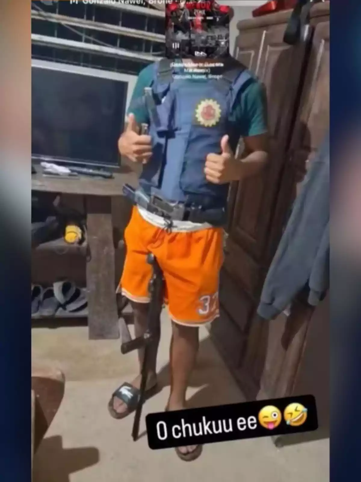 Persona con chaleco antibalas, pantalones cortos naranjas y sandalias, posando con el pulgar arriba y un arma larga apoyada en la pierna, en una habitación con muebles y una televisión. Persona con chaleco antibalas, pantalones cortos naranjas y sandalias, posando con el pulgar arriba y un arma larga apoyada en la pierna, en una habitación con muebles y una televisión.