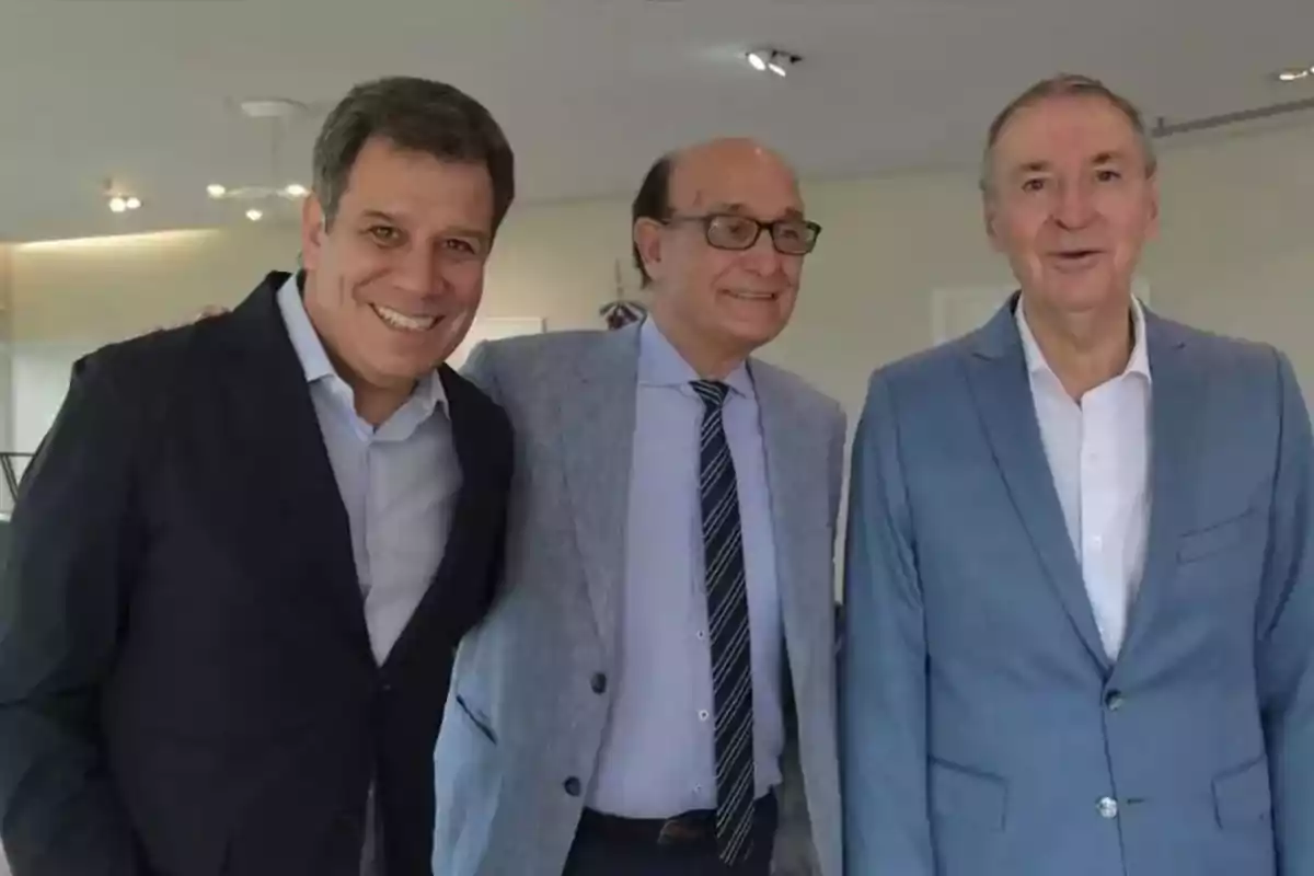 Three men dressed in jackets and jerseys pose together and smile in a well-lit indoor setting. Three men dressed in jackets and jerseys pose together and smile in a well-lit indoor setting.