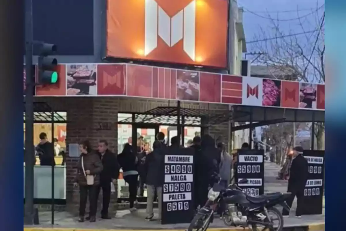 Personas haciendo fila en la entrada de una carnicería con carteles de precios y una moto estacionada en la vereda Personas haciendo fila en la entrada de una carnicería con carteles de precios y una moto estacionada en la vereda