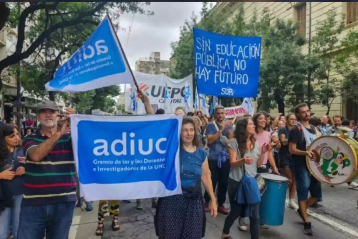 Personas marchando por la calle con pancartas y banderas en defensa de la educación pública, algunas personas tocan tambores y se observa un ambiente de protesta pacífica